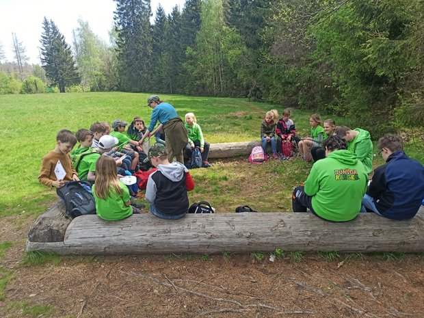 Die Junior-Ranger des Nationalparks Harz sitzen auf Baumstämmen im Kreis. Paula steht in der Mitte und erklärt das weitere Vorgehen. 