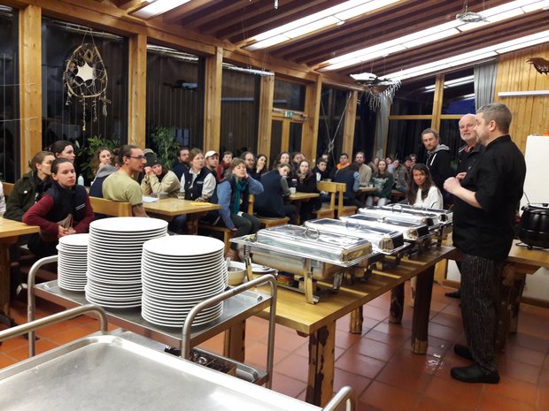 Aufgebautes Buffet mit Wärmebehältern und Menschen die den Köchen bei der Erklärung zuhören.