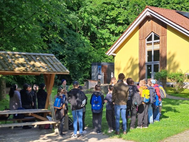 Eine Schulklasse steht im Kreis vor einem gelben Geäude und hört einer Person zu.