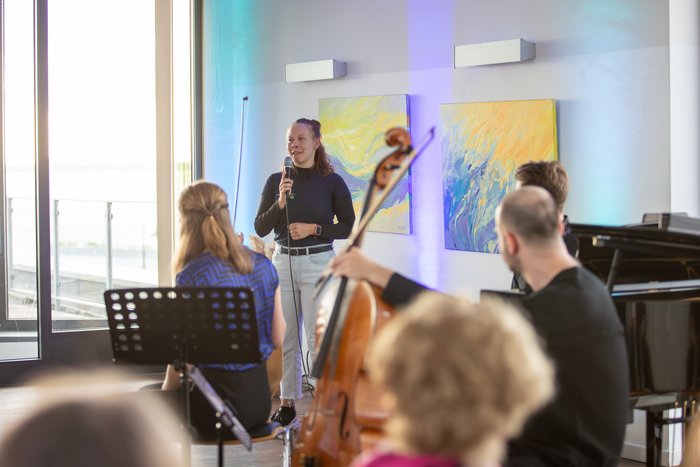Eine junge Erwachsene steht mit einem Mikrofon in einem Raum. Sie ist halb verdeckt von Musikerinnen, die vor ihr mit Notenständern und Streichinstrumenten sitzen. 