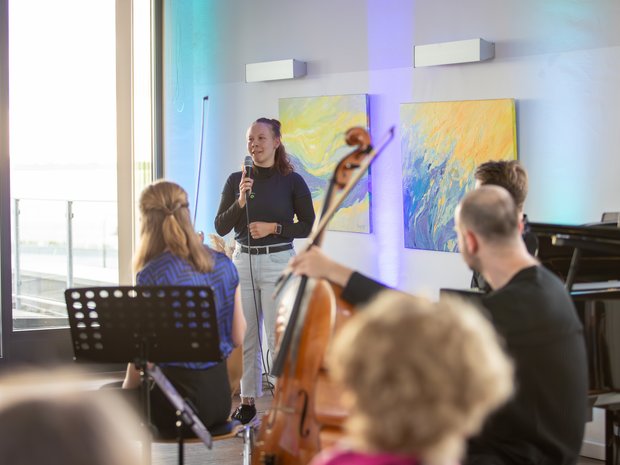 Eine junge Erwachsene steht mit einem Mikrofon in einem Raum. Sie ist halb verdeckt von Musikerinnen, die vor ihr mit Notenständern und Streichinstrumenten sitzen. 