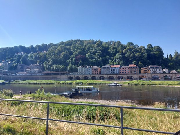 Landschaftsaufnahme mit einer anlegenden Fähre auf einem Fluss im Mittelpunkt. Im Hintergrund sind bunte, mehrstöckige Häuser, dahinter Wald zu erkennen. 