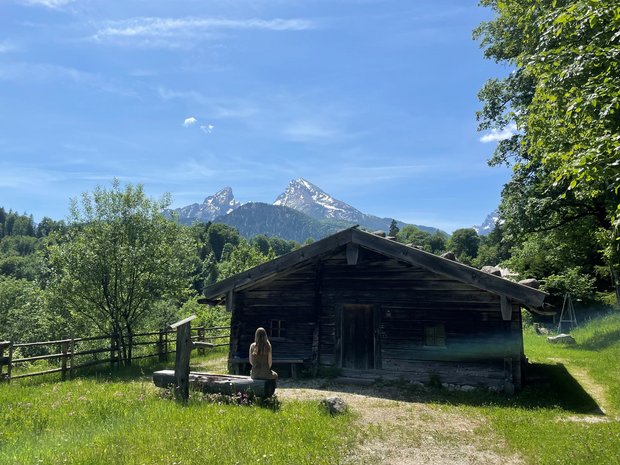 Luca Sofie sitzt auf einer Holzbank vor einer kleinen Holzhütte mit Blick auf die Berglandschaft.