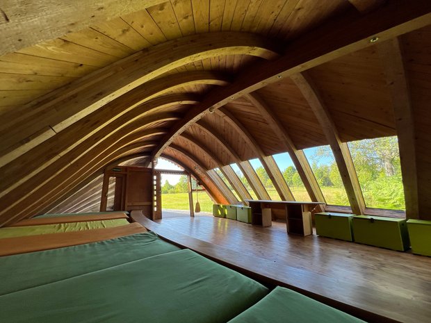 Der Innenraum einer Hütte mit Matratzenlager, geschwungenem Dach und Blick aus dem Panorama-Fenster ins Freie.