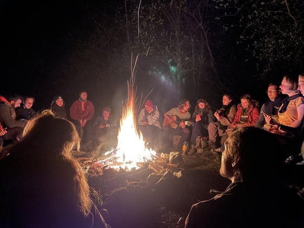 Gruppe sitzt im Kreis um ein großes Lagerfeuer herum. 