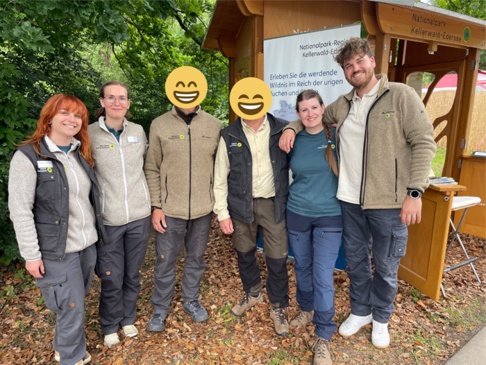 Sechs Personen stehen lachend, Arm in Arm vor einem Holzstand.