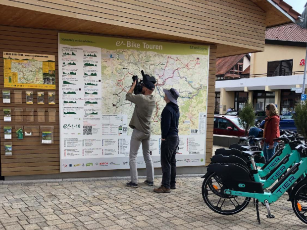 Ein Kameramann steht mit einer weiteren Person vor einer Landkarte mit E-Bike Touren. 